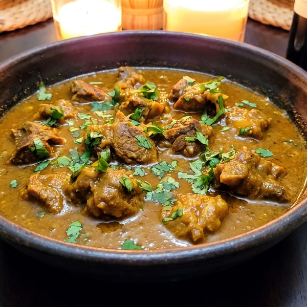 A dark bowl of rich lamb curry with fresh cilantro, wine, and candles on a woven placemat.